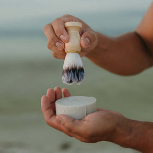 man-holds-up-tallow-shave-soap-and-shave-brush-against-nature-background