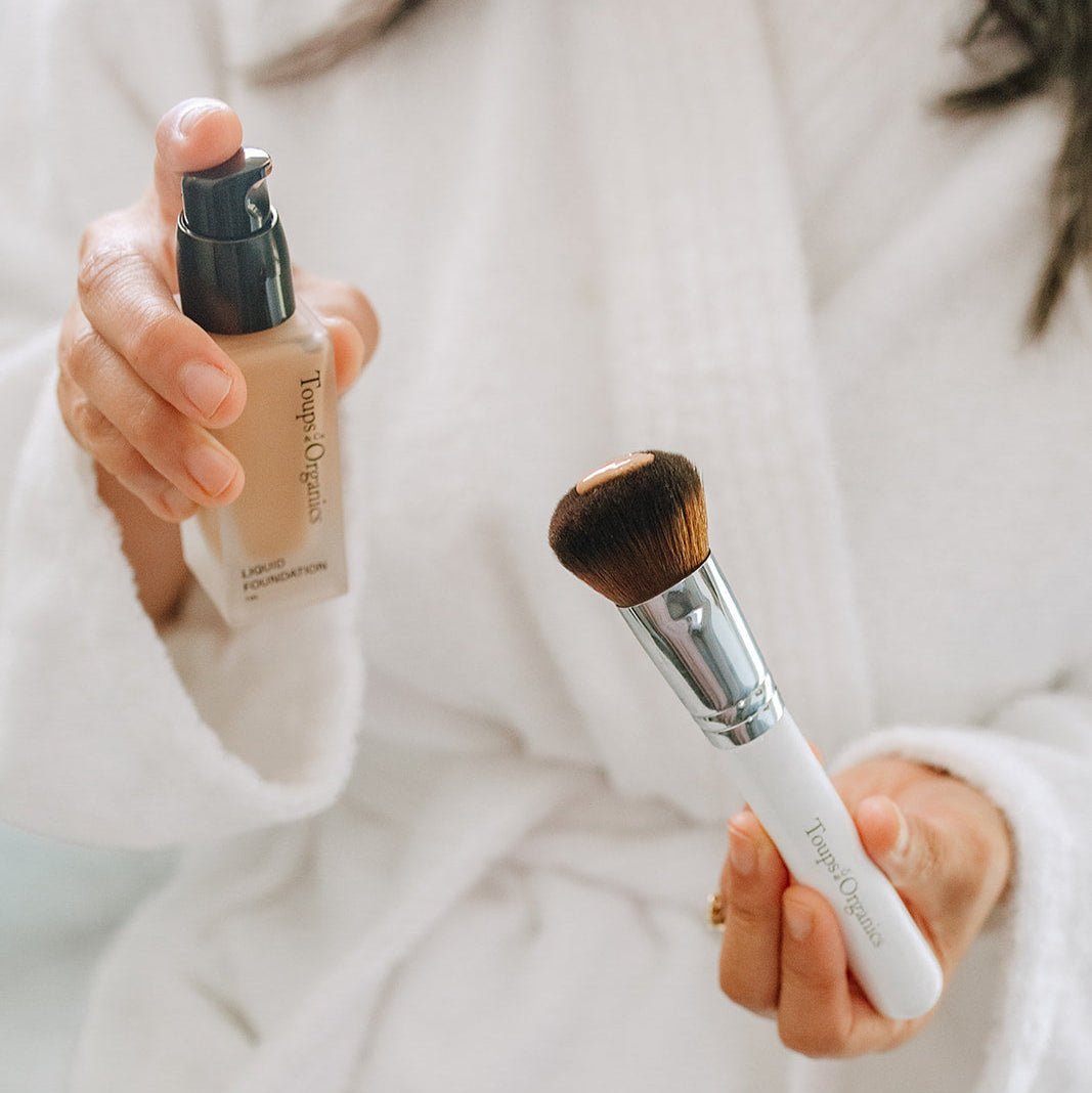 A person in a white robe holds a non-toxic foundation bottle and a Toups and Co Organics Foundation Brush.