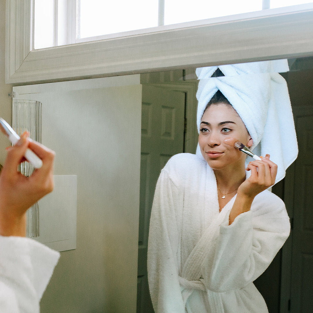 After detoxing her skin with the Toups and Co Organics Glow Face Mask, a woman in a white bathrobe and towel applies makeup while looking in the bathroom mirror.