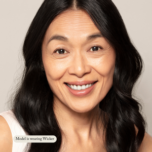 A woman with long, wavy black hair smiles at the camera, her natural glow enhanced by Toups and Co Organics Liquid Foundation for sensitive skin. Caption reads: Model is wearing Wicker.