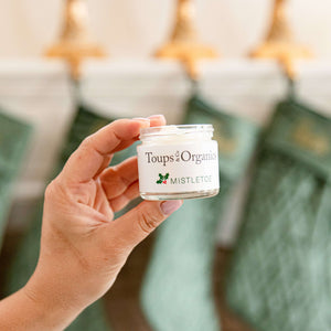 A hand holds a small jar of Tallow Balm - Mistletoe by Toups and Co Organics. Green stockings and gold hangers blur in the background—perfect for unique Christmas gifts.