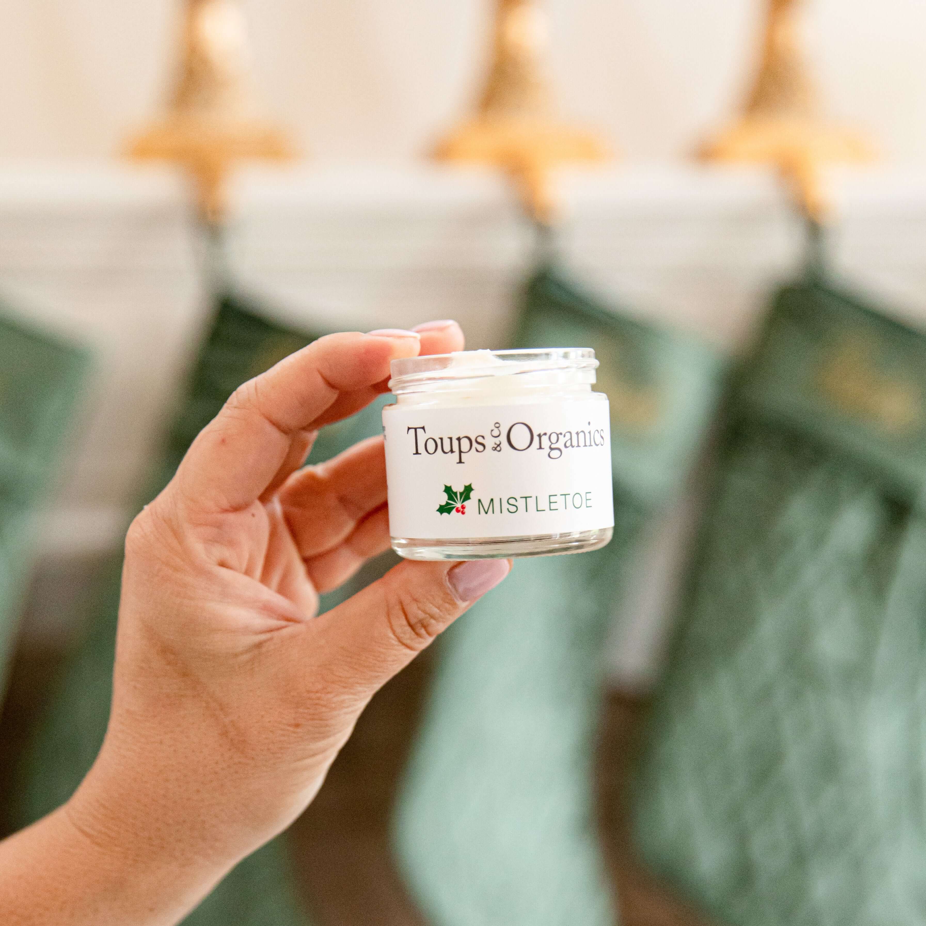 A hand holds a small jar of Tallow Balm - Mistletoe by Toups and Co Organics. Green stockings and gold hangers blur in the background—perfect for unique Christmas gifts.