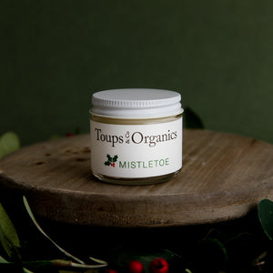 A small jar of Tallow Balm - Mistletoe by Toups and Co Organics sits on wood—ideal for Christmas gifts, with festive greenery and berries in the background.