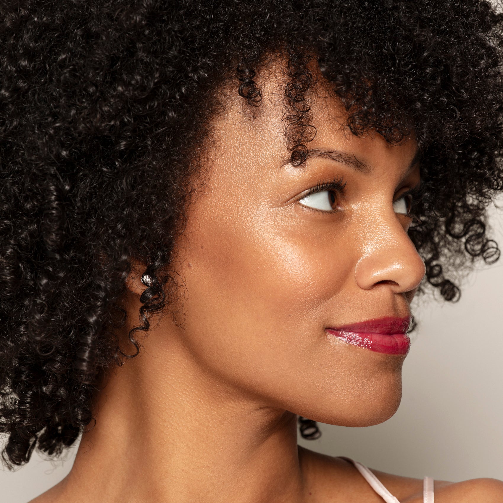 A woman with curly black hair and glossy red lipstick looks to the right against a neutral background, her complexion radiant from Toups and Co Organics Bronzer for a subtle sun-kissed glow.
