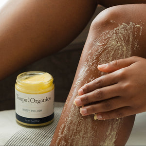 A person applies Toups and Co Organics Body Polish to their leg, with the open jar beside them.