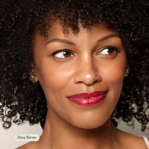 A person with curly dark hair, in glossy red lipstick, smiles softly. A label reads Deep Brown—Toups and Co Organics Brow Pencil, ideal for smooth, buttery application and perfect eyebrow color.