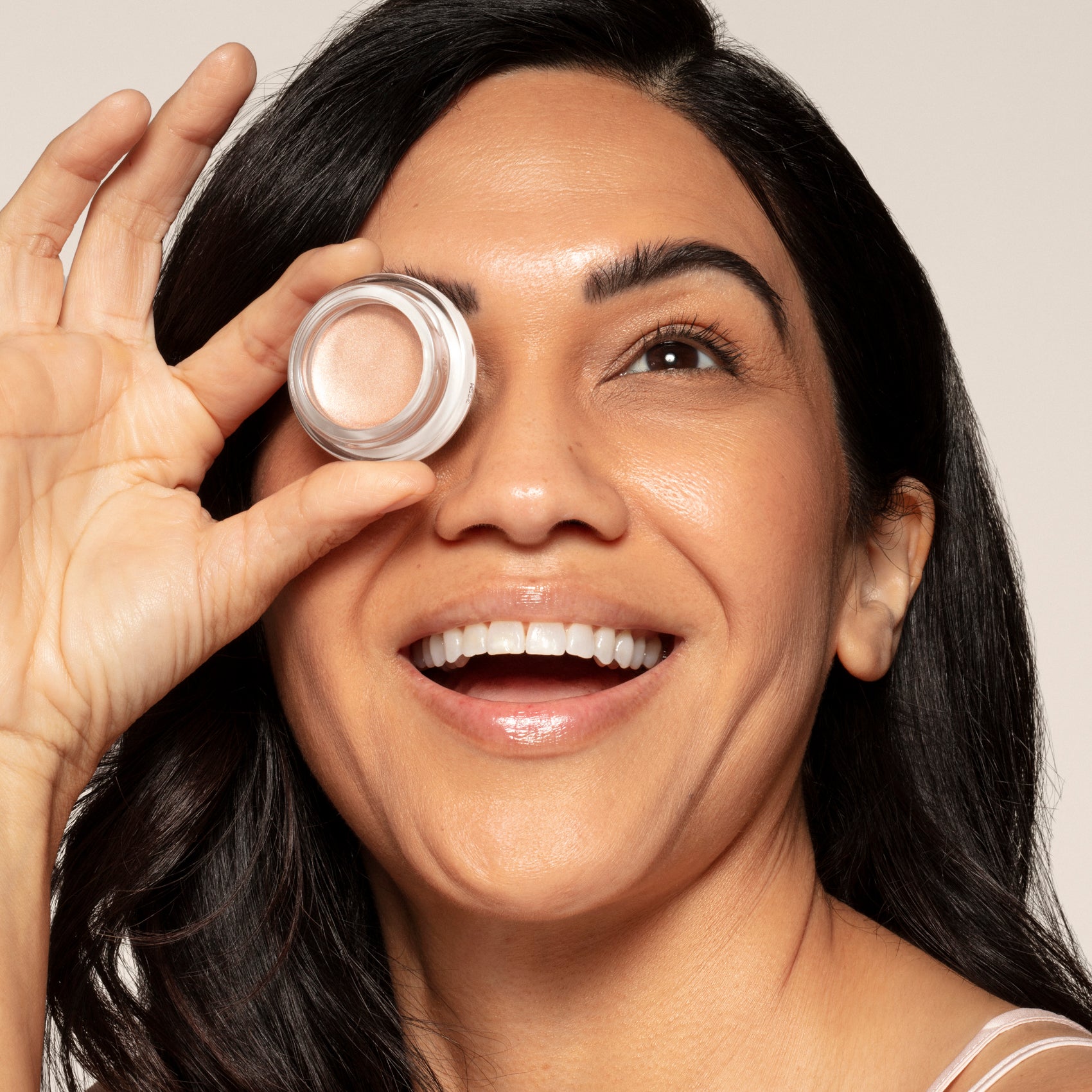 A smiling woman holds a small round Toups and Co Organics Cream Luminizer Pot up to one eye, her mouth slightly open and teeth visible, against a plain background.