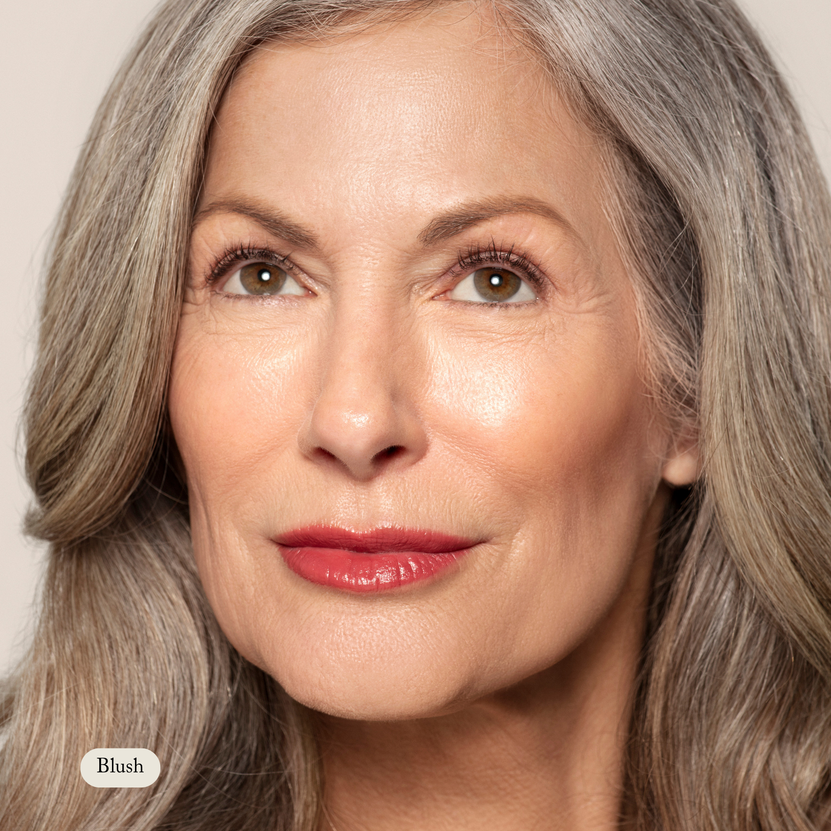 Close-up of an older woman with long gray hair and red lipstick, looking up and slightly right, showcasing a natural glow from Toups and Co Organics Cream Cheek Tint against a neutral background.