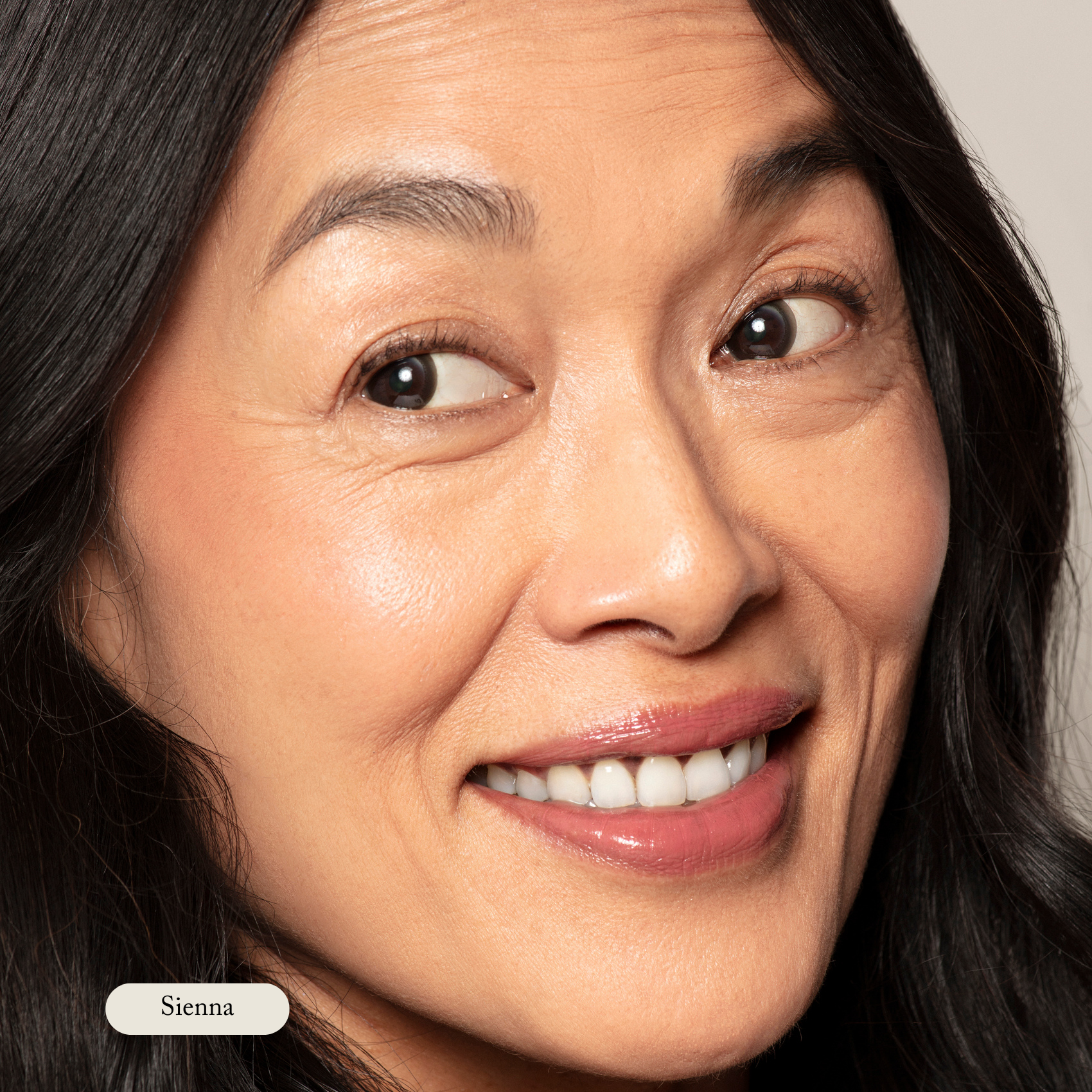 Close-up of a smiling woman with long black hair and smooth skin, wearing Toups and Co Organics Cream Cheek Tint in Sienna. The natural glow of her cheeks is enhanced by the tint. A small label at the bottom left reads Sienna.