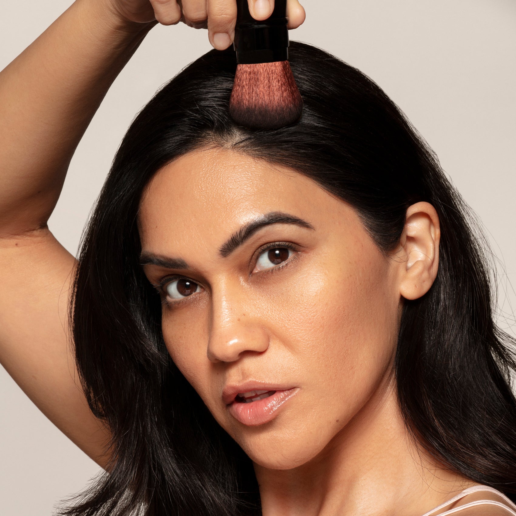 A woman applies Toups and Co Organics Dry Shampoo to her hairline with a makeup brush, demonstrating dark color options for brown hair while looking directly at the camera.