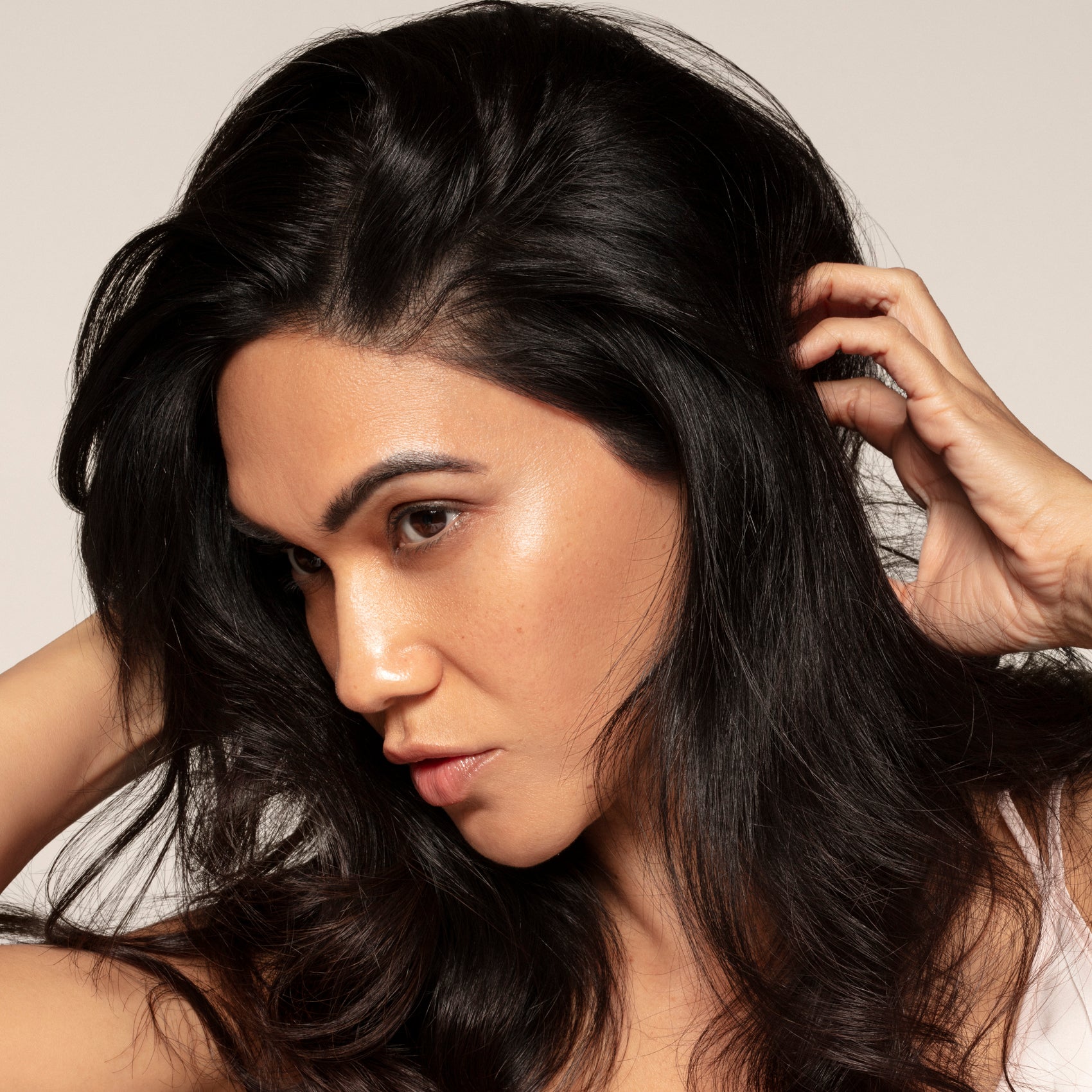 A woman with long dark hair touches her head and looks to the side, perhaps considering Toups and Co Organics Dry Shampoo for brown hair, against a plain background.