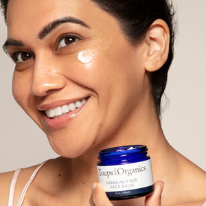A woman smiles as she applies Toups and Co Organics Frankincense Face Balm, holding the blue jar of this nourishing, regenerative moisturizer near her cheek.