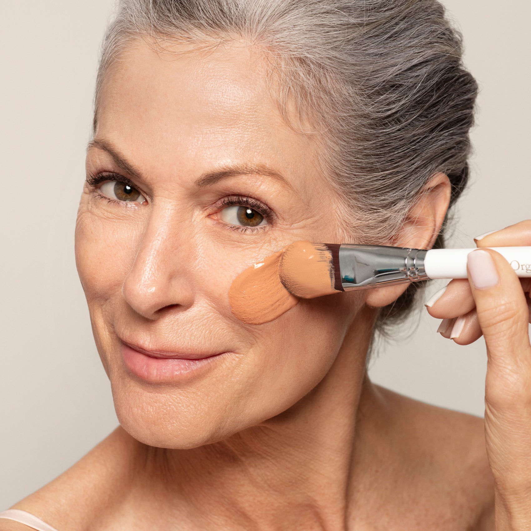 An older woman with gray hair smiles as she applies liquid foundation, her radiant skin showing the detoxifying benefits of Toups and Co Organics Glow Face Mask—her favorite skincare essential.