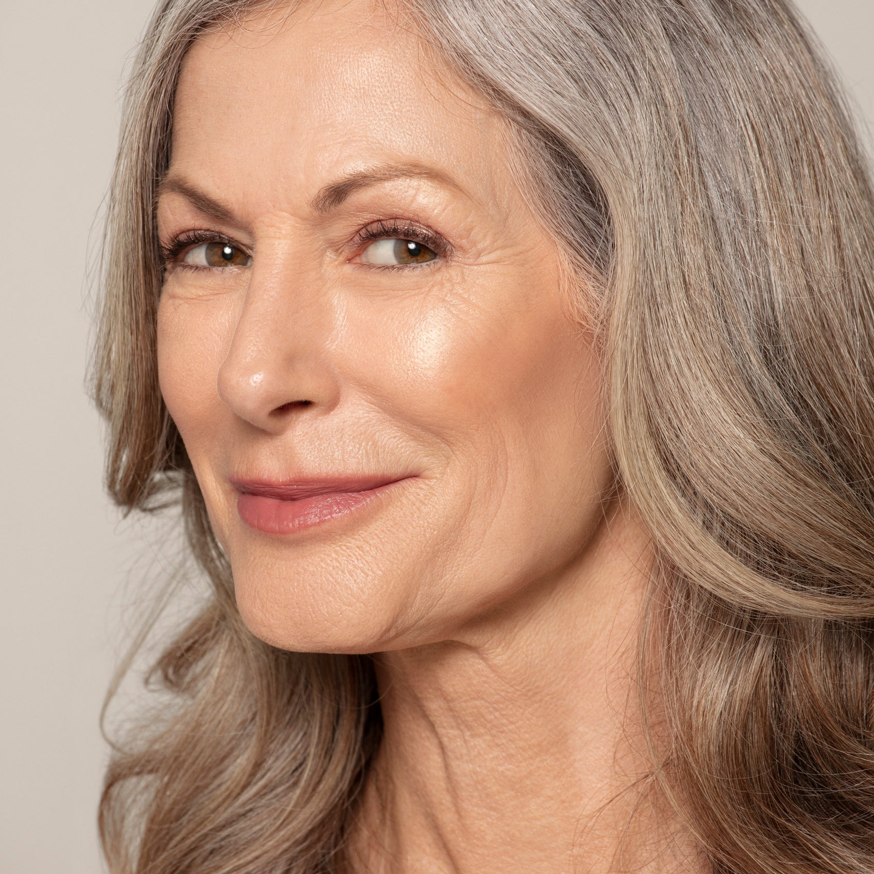 Close-up of an older woman with long gray hair and light makeup, smiling subtly as her skin glows from Toups and Co Organics Highlighter, an all-natural formula with brilliant pigmentation.