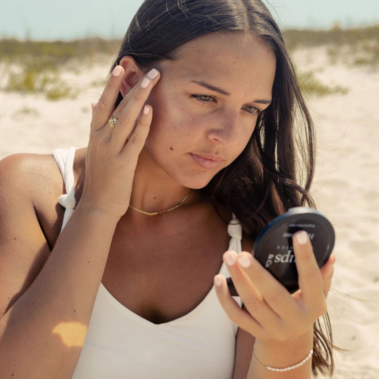 A woman at the beach applying product to her cheek while looking into a handheld mirror.