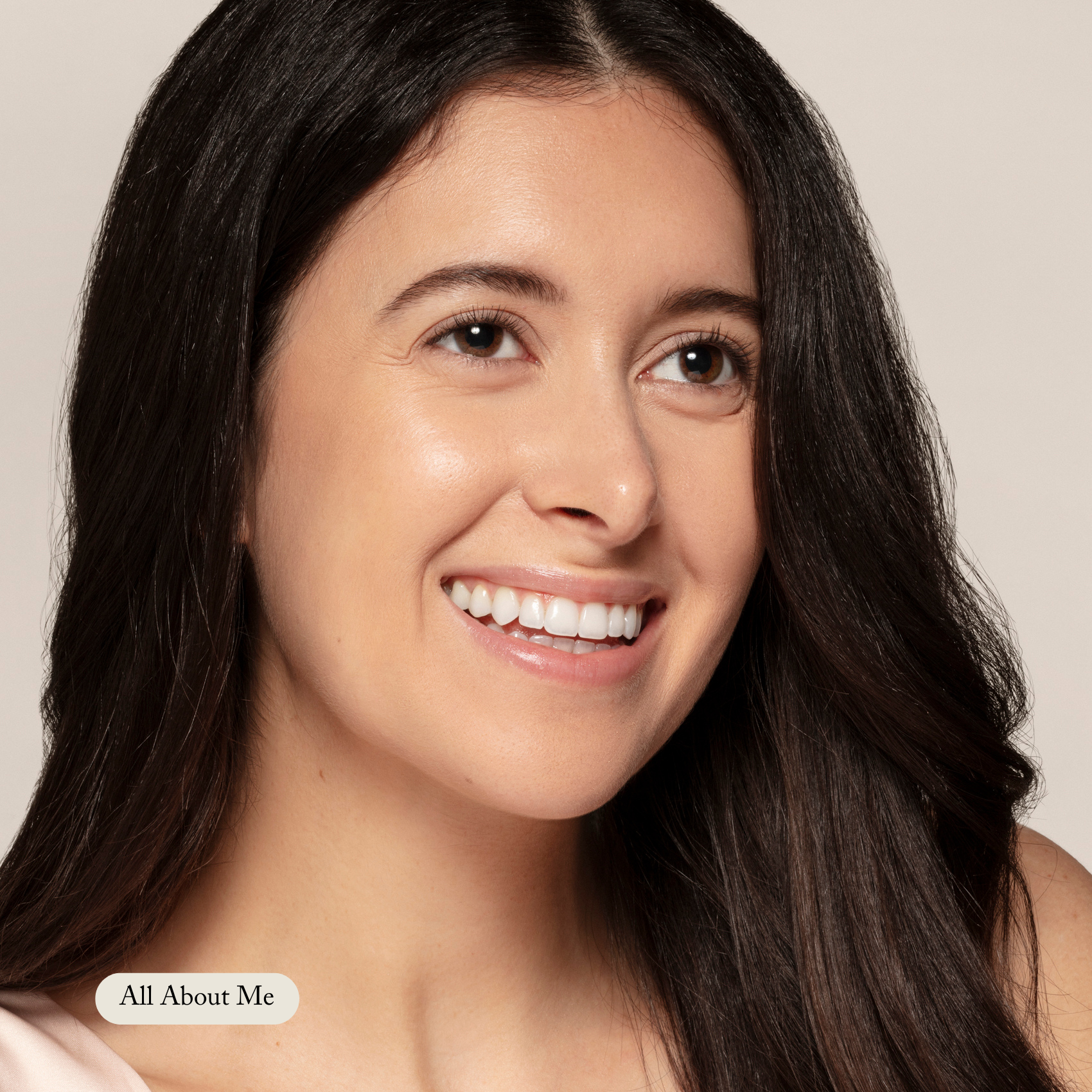 A woman with long dark hair smiles while looking off-camera. A neutral background highlights her radiant look, and a button labeled All About Me in the lower left corner showcases Toups and Co Organics’ Organic Lip Gloss.