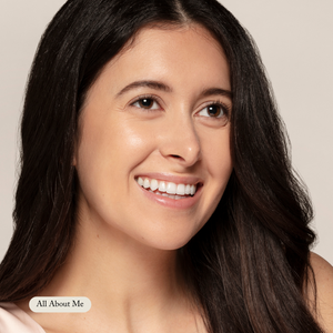 A woman with long dark hair smiles while looking off-camera. A neutral background highlights her radiant look, and a button labeled All About Me in the lower left corner showcases Toups and Co Organics’ Organic Lip Gloss.