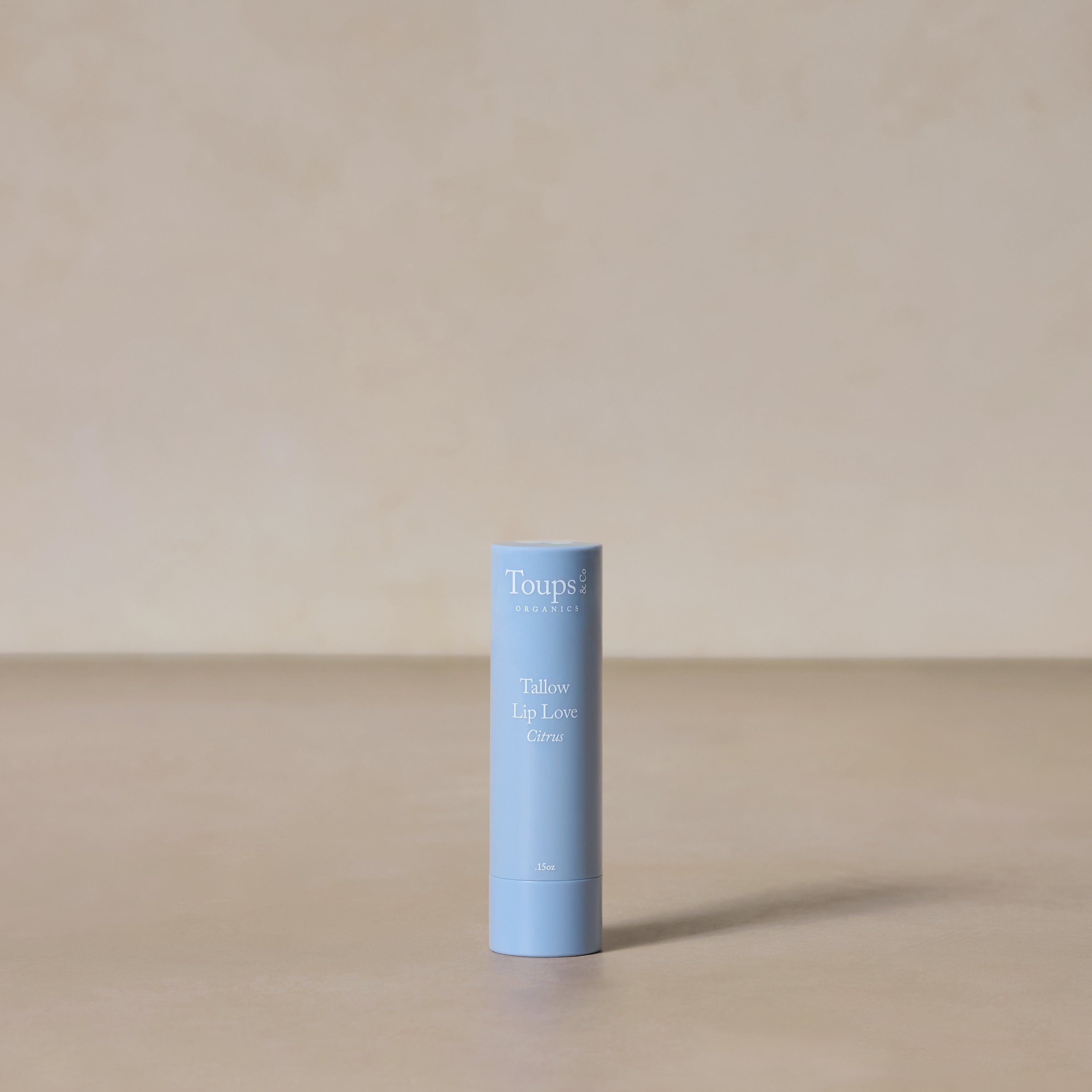 A light blue tube of Toups and Co Organics Tallow Lip Love stands upright on a neutral beige surface with a matching background—ideal for soothing chapped lips.