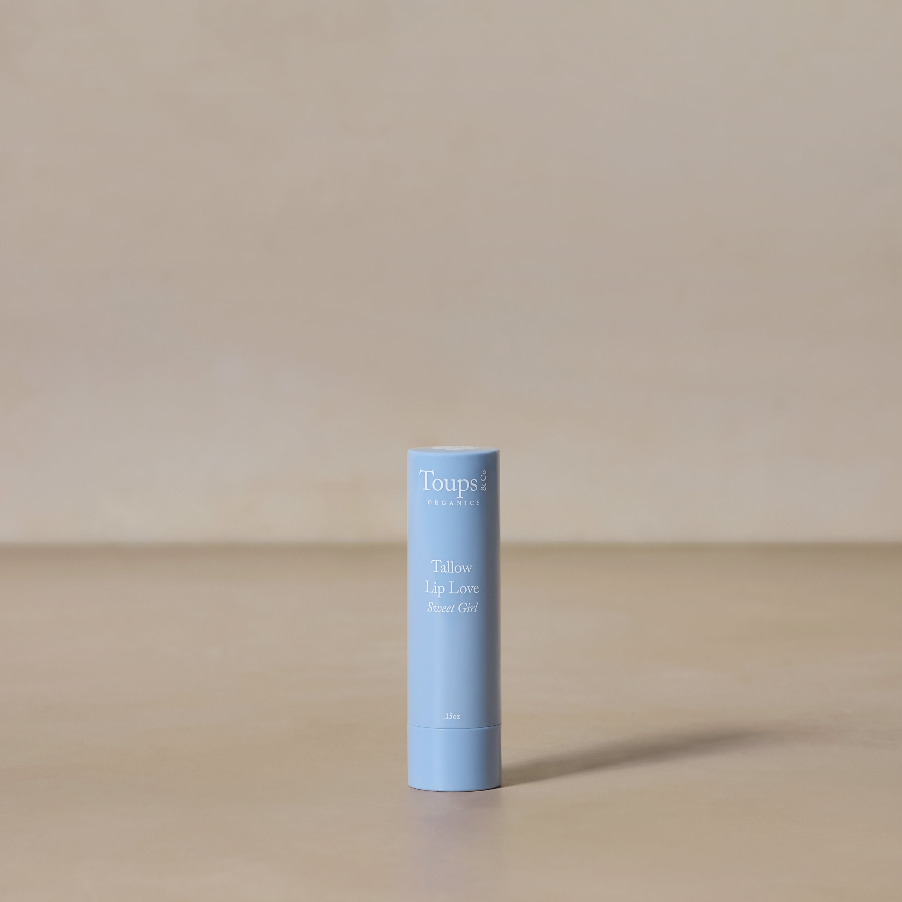 A light blue tube of Toups and Co Organics Tallow Lip Love hydrating lip balm stands upright on a beige surface with a neutral background.