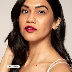 A woman with long dark hair wears Toups and Co Organics Nourishing Lipstick in Black Cherry, featuring hydrating butters and antioxidants, paired with a white strap top as she looks at the camera.