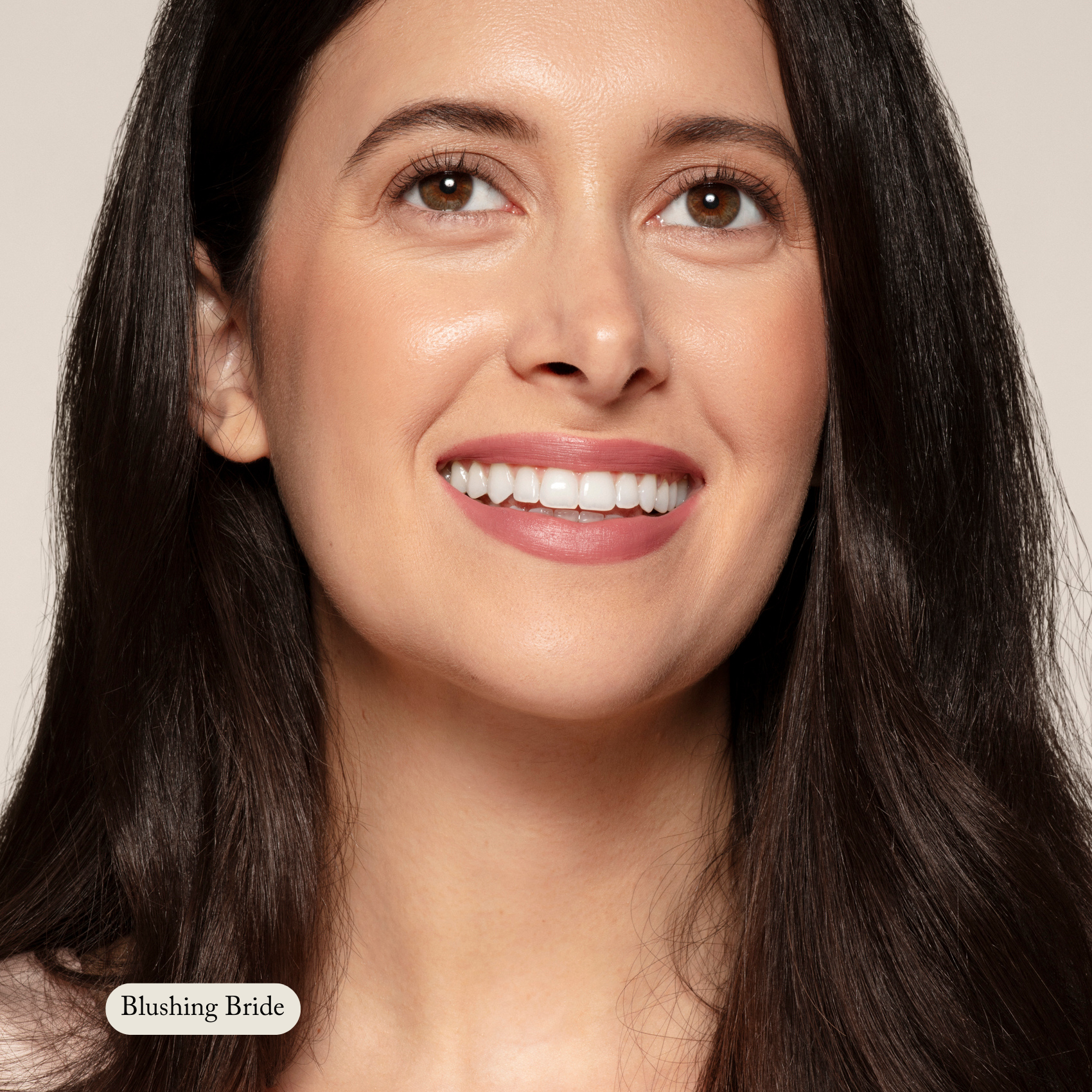 A woman with long dark hair and clear skin smiles, wearing Toups and Co Organics Nourishing Lipstick in Blushing Bride—a hydrating, buttery formula giving her lips a soft, healthy look with subtle pink color.