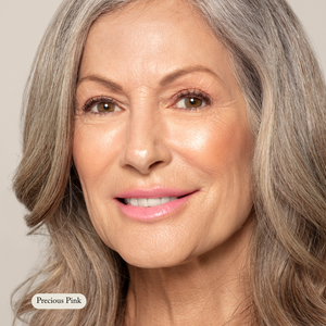 A woman with long gray hair smiles, wearing Toups and Co Organics Nourishing Lipstick in Precious Pink—infused with hydrating butters and antioxidants.