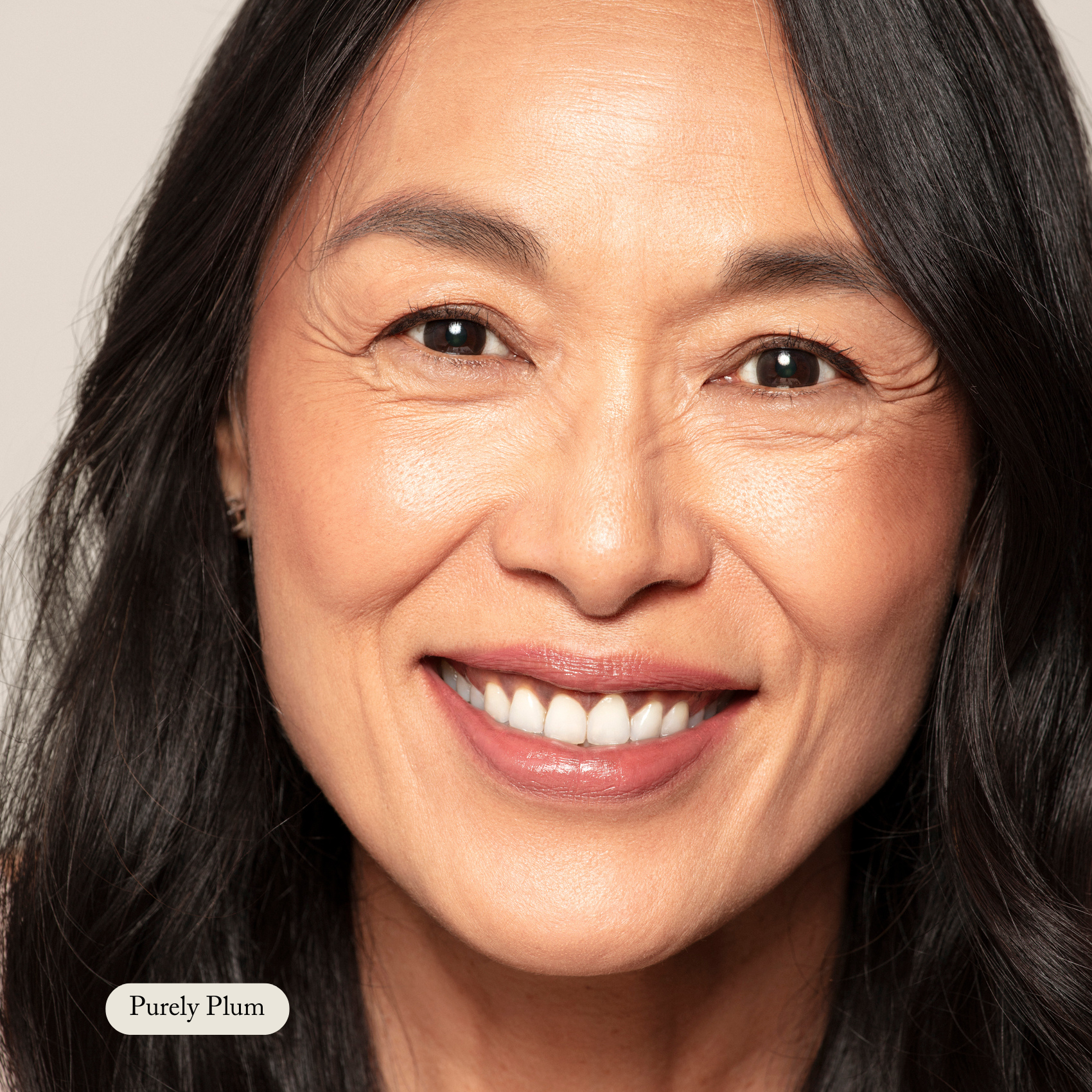 A middle-aged woman with long dark hair smiles at the camera against a plain background. A small label reads Purely Plum, highlighting her look enhanced by Toups and Co Organics Nourishing Lipstick.