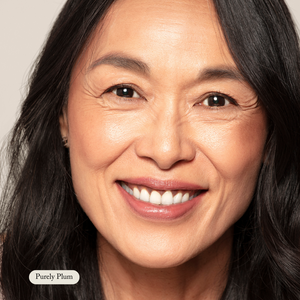 A middle-aged woman with long dark hair smiles at the camera against a plain background. A small label reads Purely Plum, highlighting her look enhanced by Toups and Co Organics Nourishing Lipstick.