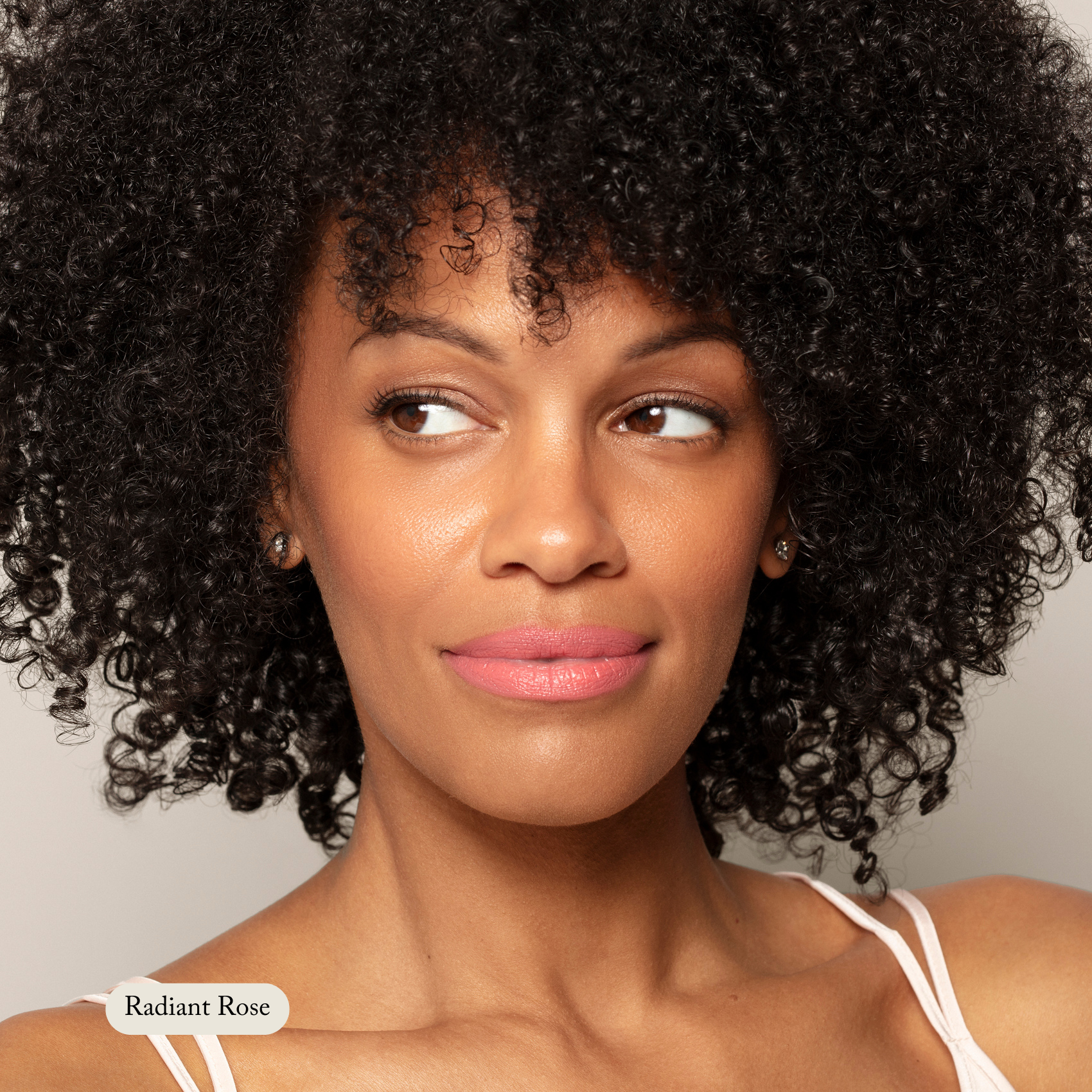 A woman with curly black hair and radiant skin glances sideways, wearing Toups and Co Organics’ Nourishing Lipstick in Radiant Rose, enriched with hydrating butters for moisture. She has on a white strap.