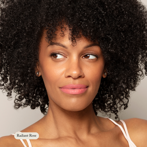 A woman with curly black hair and radiant skin glances sideways, wearing Toups and Co Organics’ Nourishing Lipstick in Radiant Rose, enriched with hydrating butters for moisture. She has on a white strap.
