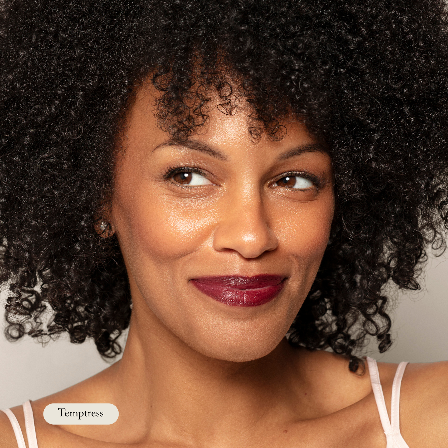 A woman with curly hair softly smiles, wearing Toups and Co Organics Nourishing Lipstick in Temptress—a bold dark red shade infused with hydrating butters—and small stud earrings.