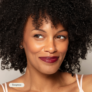 A woman with curly hair softly smiles, wearing Toups and Co Organics Nourishing Lipstick in Temptress—a bold dark red shade infused with hydrating butters—and small stud earrings.