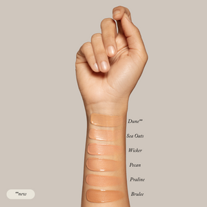 A hand displays six Toups and Co Organics Liquid Foundation swatches—Dune (new), Sea Oats, Wicker, Pecan, Praline, Brulee—on the inner forearm for a natural glow on sensitive skin, set against a neutral background.