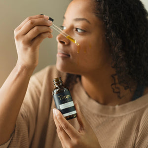 A person applying Toups Organics Seabuckthorn Cleansing Oil from a dropper bottle onto their cheek.