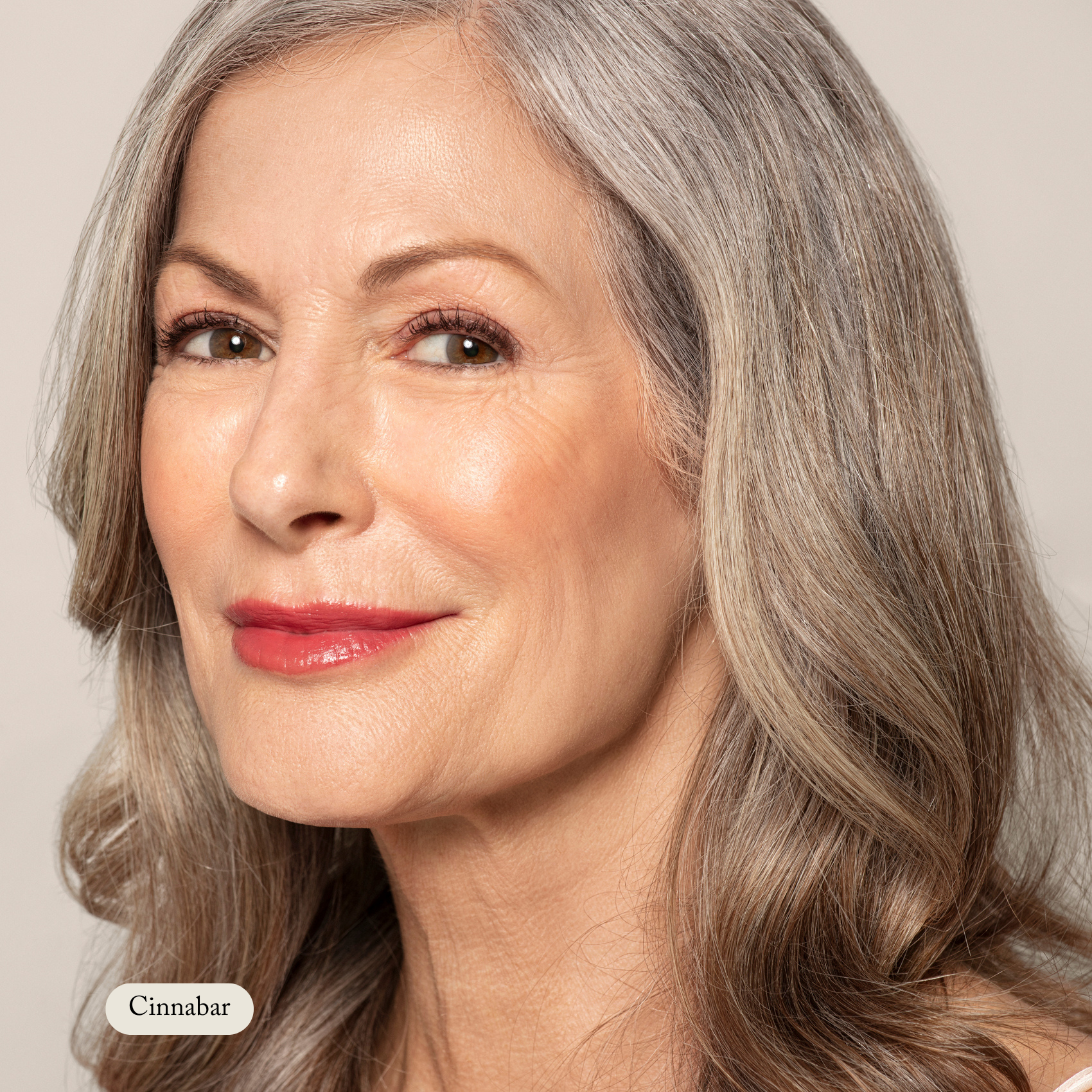 An older woman with long gray hair and red lipstick smiles at the camera against a plain backdrop. A label reads Natural Pressed Blush by Toups and Co Organics, hinting at the highly pigmented blush made with natural ingredients.