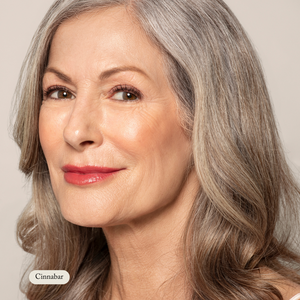 An older woman with long gray hair and red lipstick smiles at the camera against a plain backdrop. A label reads Natural Pressed Blush by Toups and Co Organics, hinting at the highly pigmented blush made with natural ingredients.