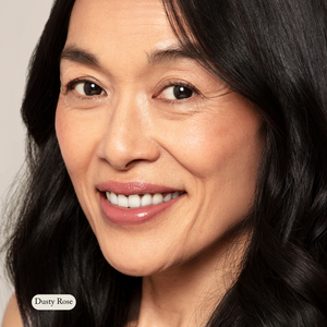 A woman with long black hair smiles, wearing natural makeup featuring Toups and Co Organics’ Natural Pressed Blush—ideal for sensitive skin—and a pinkish nude lipstick labeled Dusty Rose.