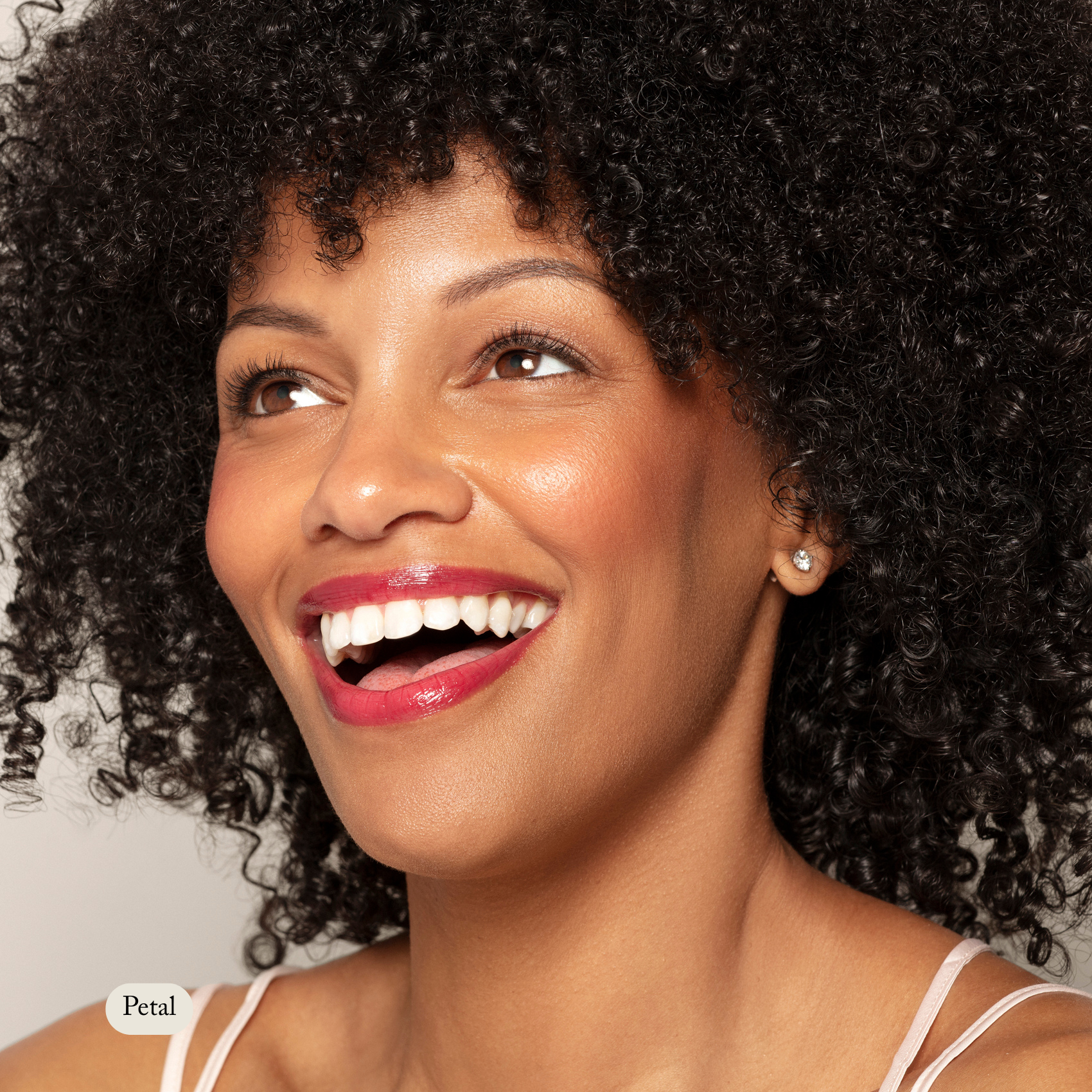 A curly-haired woman with glossy red lipstick smiles upward, her cheeks glowing with Toups and Co Organics Natural Pressed Blush, while wearing a sleeveless top and stud earrings.