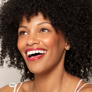 A curly-haired woman with glossy red lipstick smiles upward, her cheeks glowing with Toups and Co Organics Natural Pressed Blush, while wearing a sleeveless top and stud earrings.