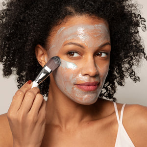 A woman in a white camisole applies Toups and Co Organics Resurfacing Mask with blue C-phycocyanin algae extract to her cheek using a brush, looking to the side.