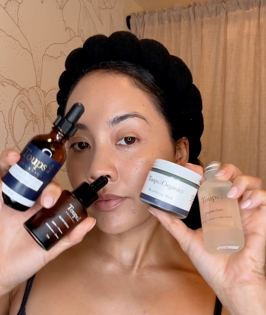 A person wearing a black headband holds four Toups and Co. skincare products: two dropper bottles, a jar of resurfacing mask, and a bottle of brightening tonic.