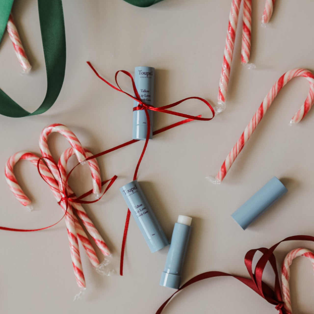 Candy Cane Lip Love lip balm tubes by Toups and Co Organics, tied with red ribbons, are displayed with candy canes and green and red decorative ribbon on a beige surface.