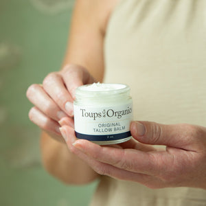 A person holds an open jar of Tallow Balm - Original by Toups and Co Organics, with a dab on their fingers—perfect for full-body moisture and sensitive, dry skin.