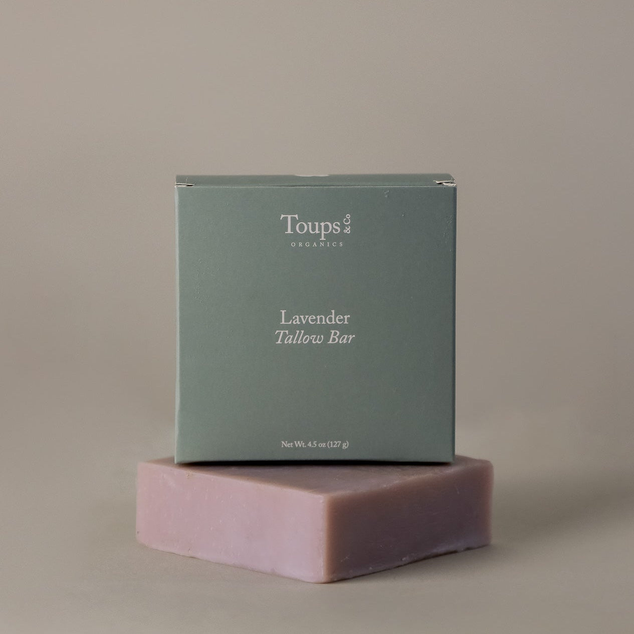 A green box labeled Toups and Co Organics Lavender Tallow Bar rests atop a lavender-colored bar of soap made with grass-fed tallow, set against a neutral background to showcase this natural dry skin solution.