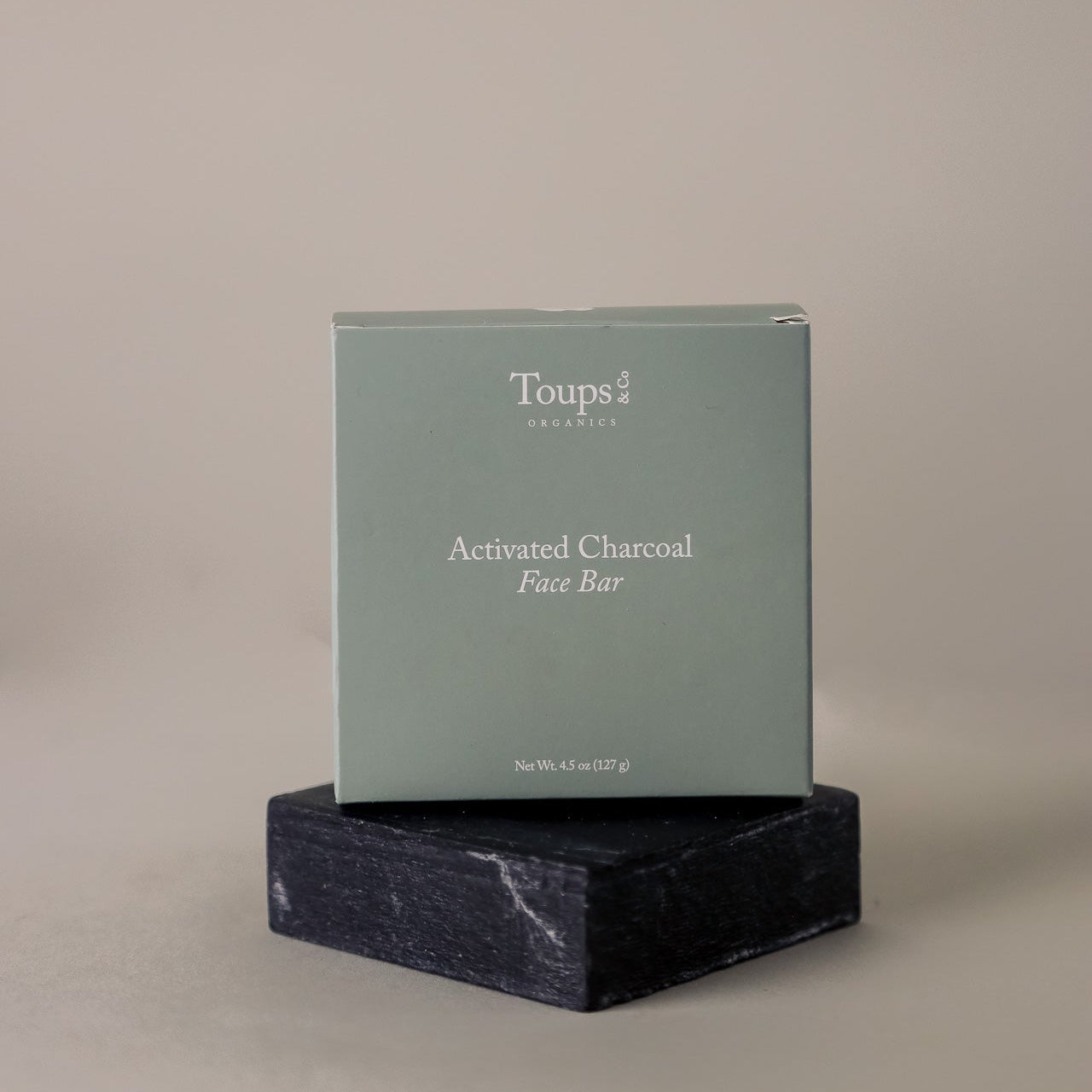 A box labeled Toups and Co Organics Activated Charcoal Face Bar, made with grass-fed tallow and activated bamboo charcoal, sits on a dark stone platform against a plain background.