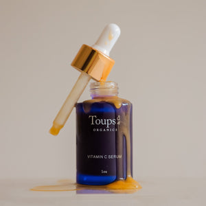 A glass bottle of Toups and Co Organics Vitamin C Serum features a dropper, with serum spilling and dripping down the bottle onto the surface.