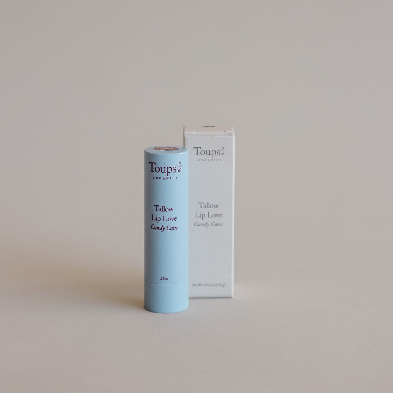 A light blue tube of Toups and Co Organics Candy Cane Lip Love, enriched with grass-fed tallow, stands beside its white box packaging on a neutral background.