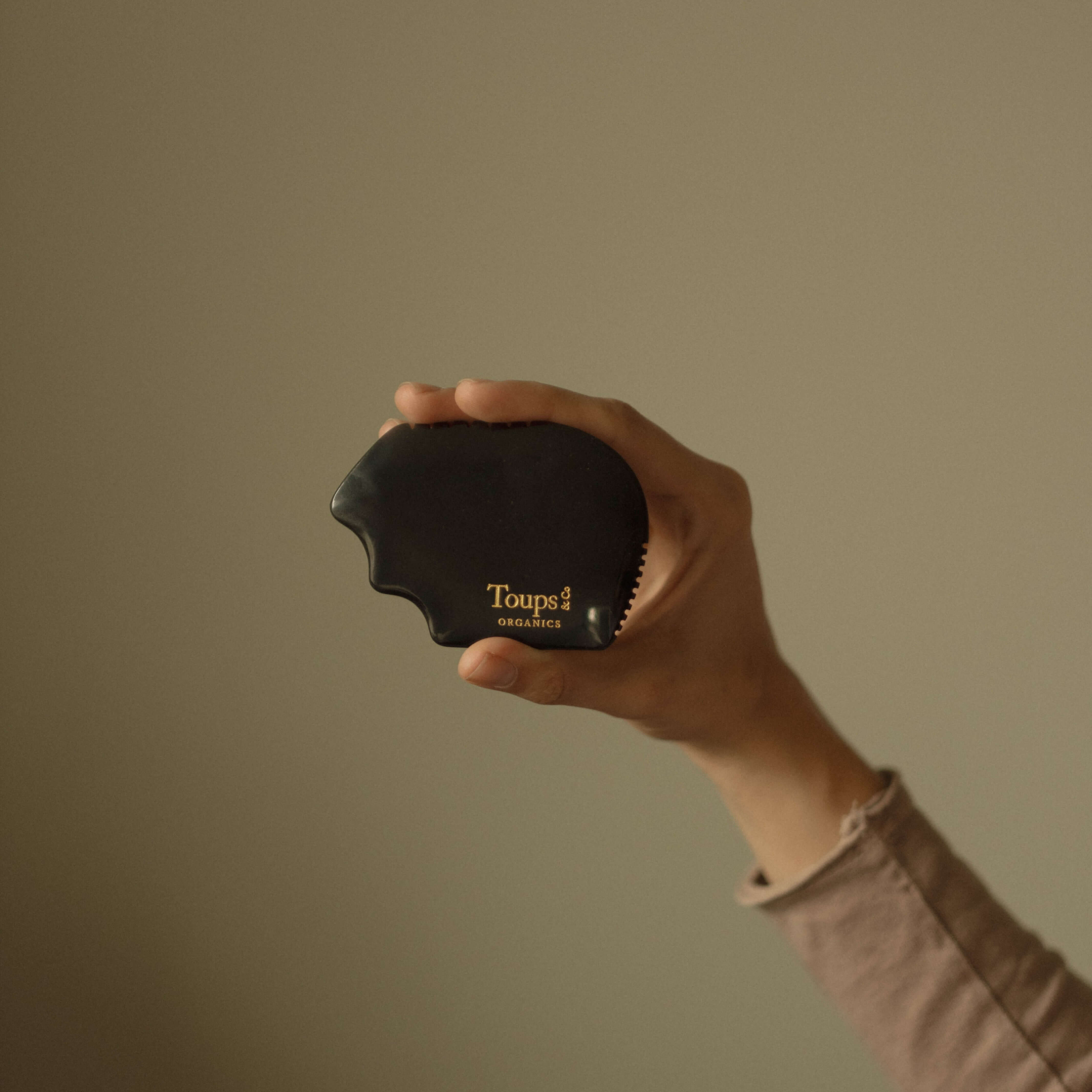 A hand holds a black Nephrite Gua Sha by Toups and Co Organics, ideal for lymphatic drainage, set against a plain beige background.