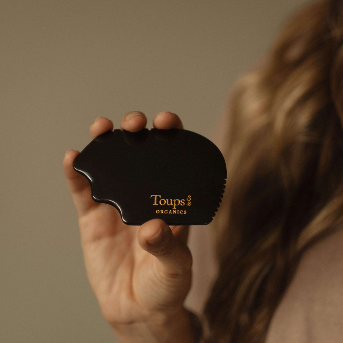 A hand holds the Nephrite Gua Sha by Toups and Co Organics, featuring gold lettering, ideal for lymphatic drainage massage against a subtle background.