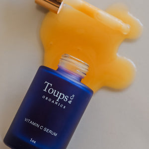 A blue bottle of Toups and Co Organics Vitamin C Serum lies on its side, with some serum spilled onto a light surface.